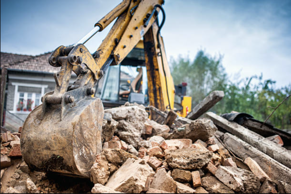 demolition-bunker-hill-tree A bulldozer is clearing rubble in front of a house, preparing the site for construction or renovation work.